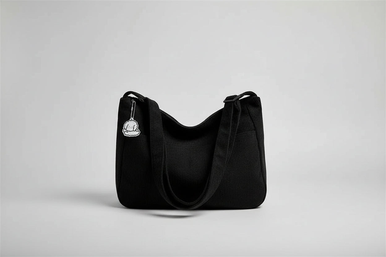 Black corduroy handbag with a keychain on a white surface near a window with blinds.
